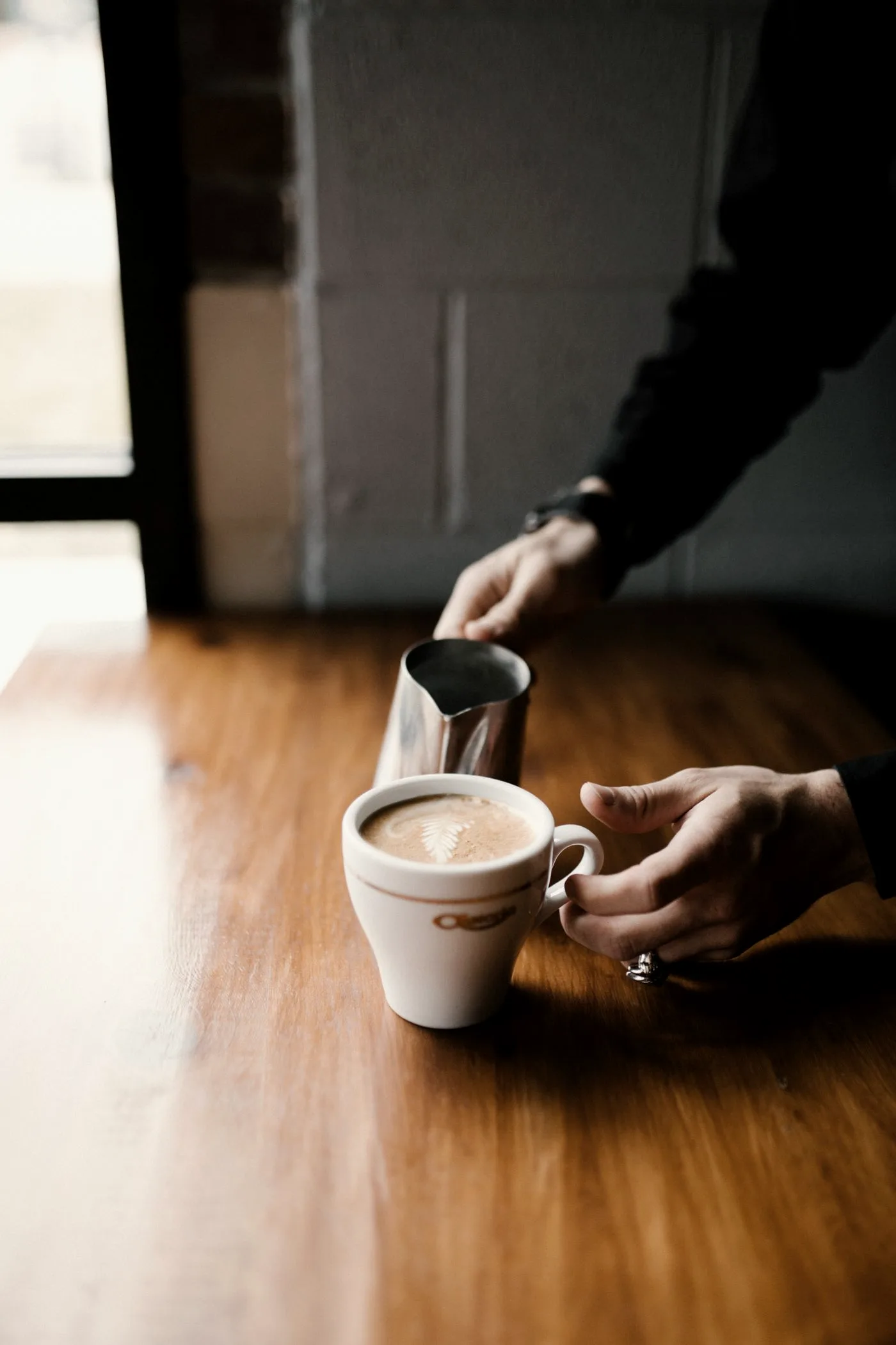 Discover Our Story Barista pouring steamed milk into a cup of coffee on a wooden café counter