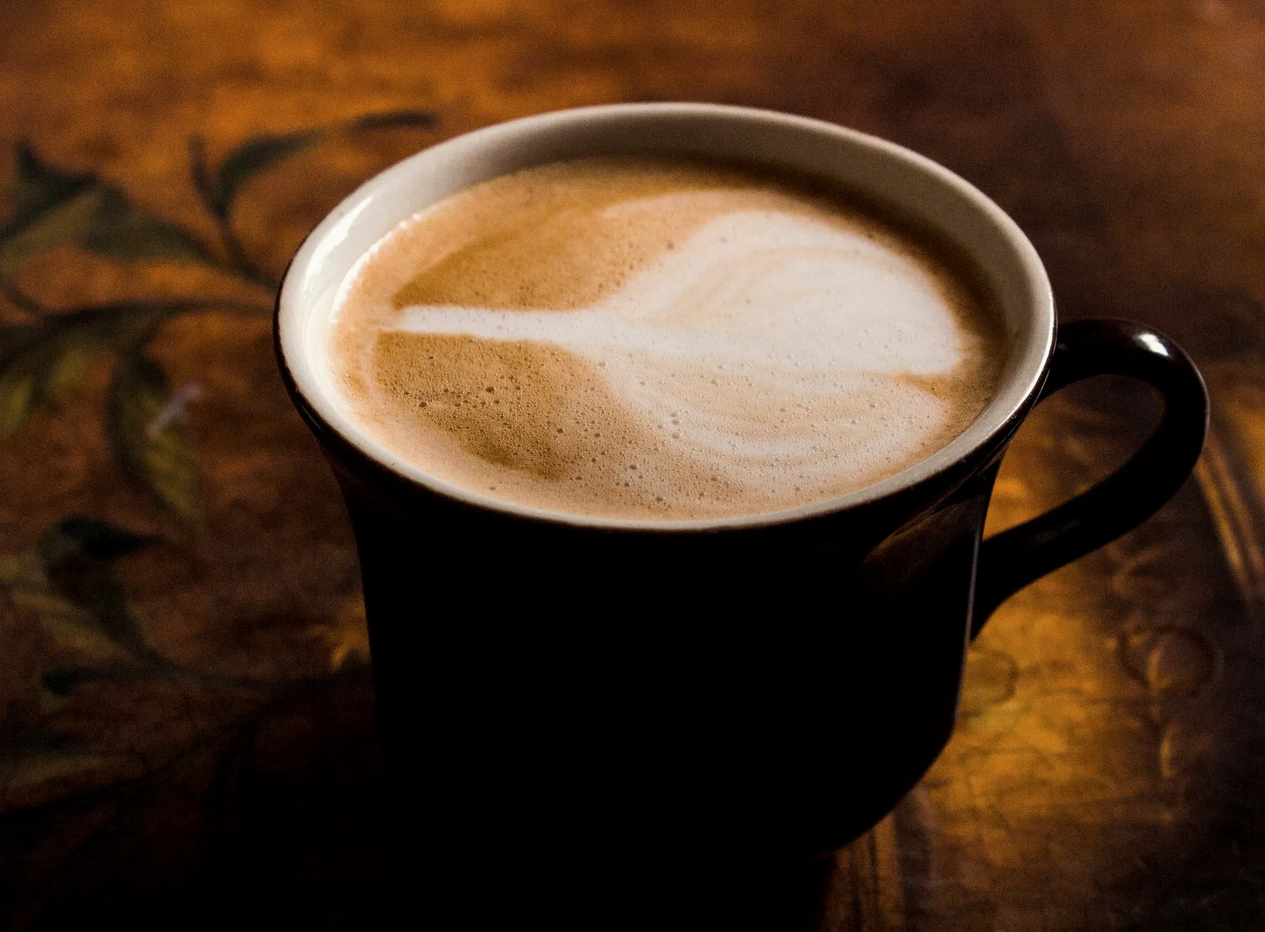 Discover Our Story Cappuccino with latte art in a dark ceramic mug on a warm wooden table
